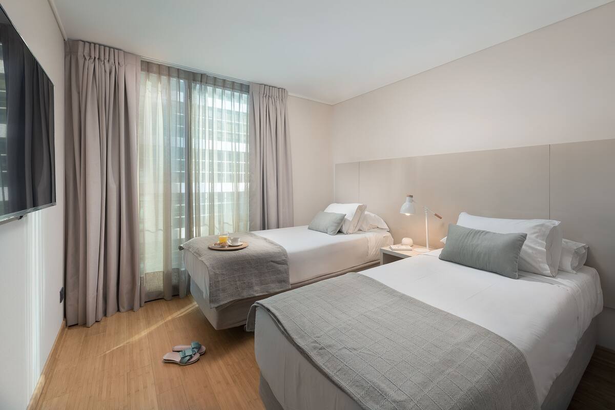 A hotel room with two beds, a TV, a bedside table with a lamp, and a window with curtains.