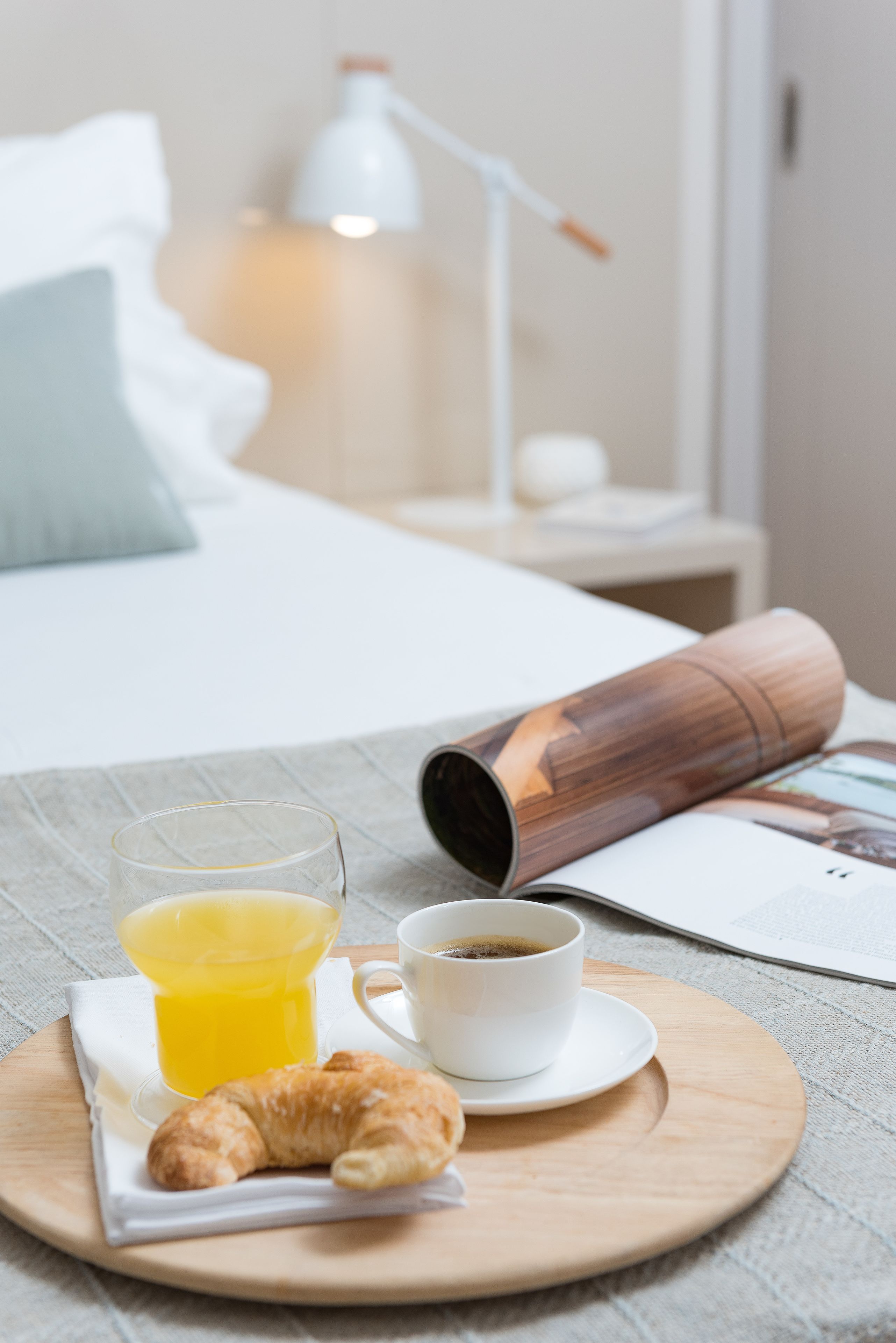 A breakfast tray with croissant, orange juice, and coffee on a wooden board, a magazine, and a lamp in the background.