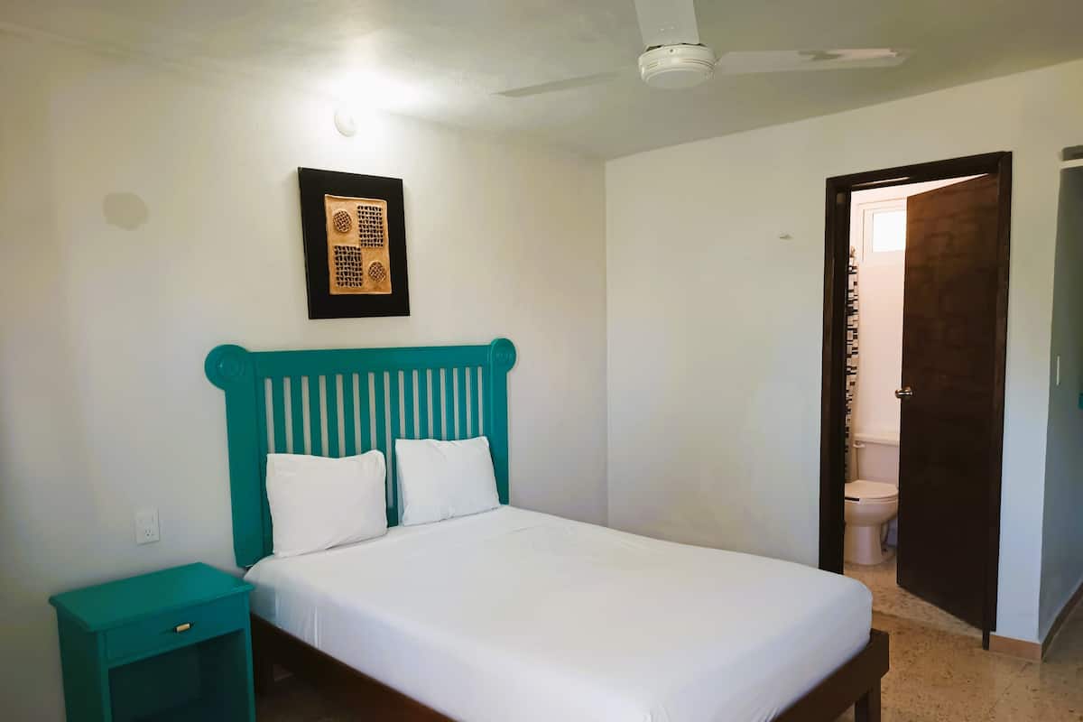 Une chambre à coucher comprenant un lit, une table de chevet, un ventilateur de plafond et une porte donnant accès à une salle de bain.