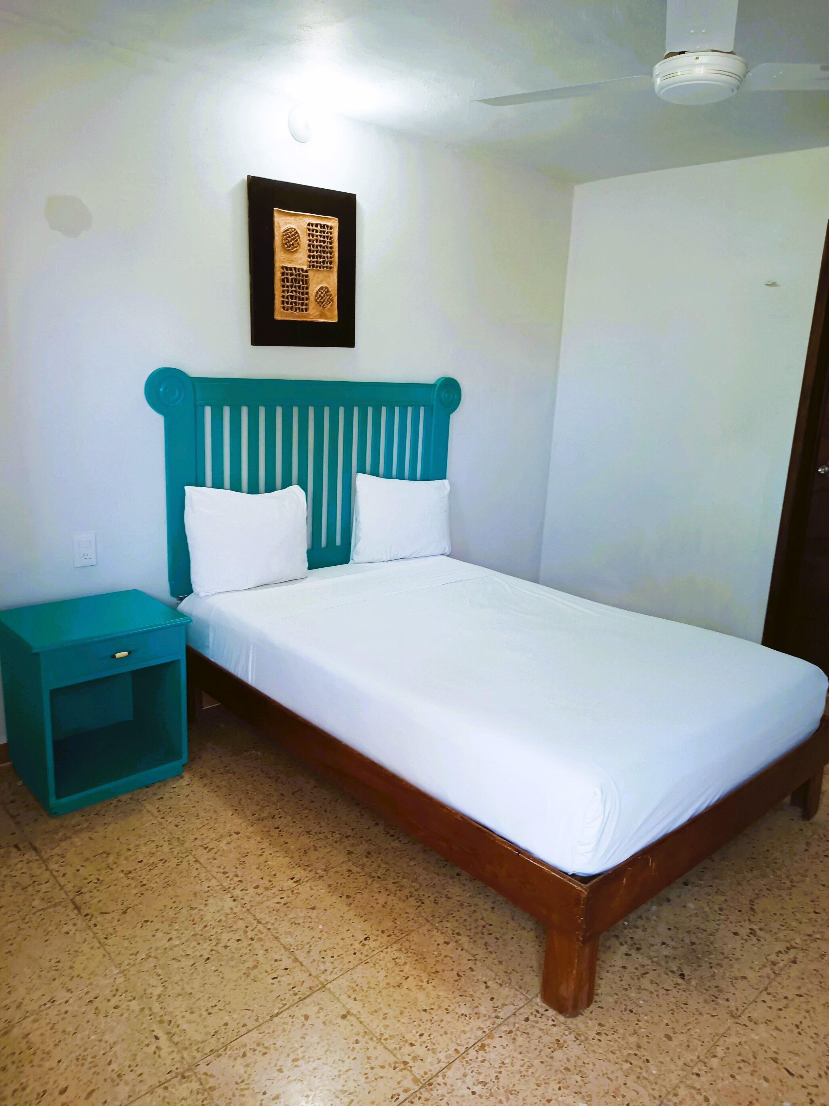 Un dormitorio con una mesita de noche verde azulado, una cama con ropa de cama blanca y un ventilador de techo.