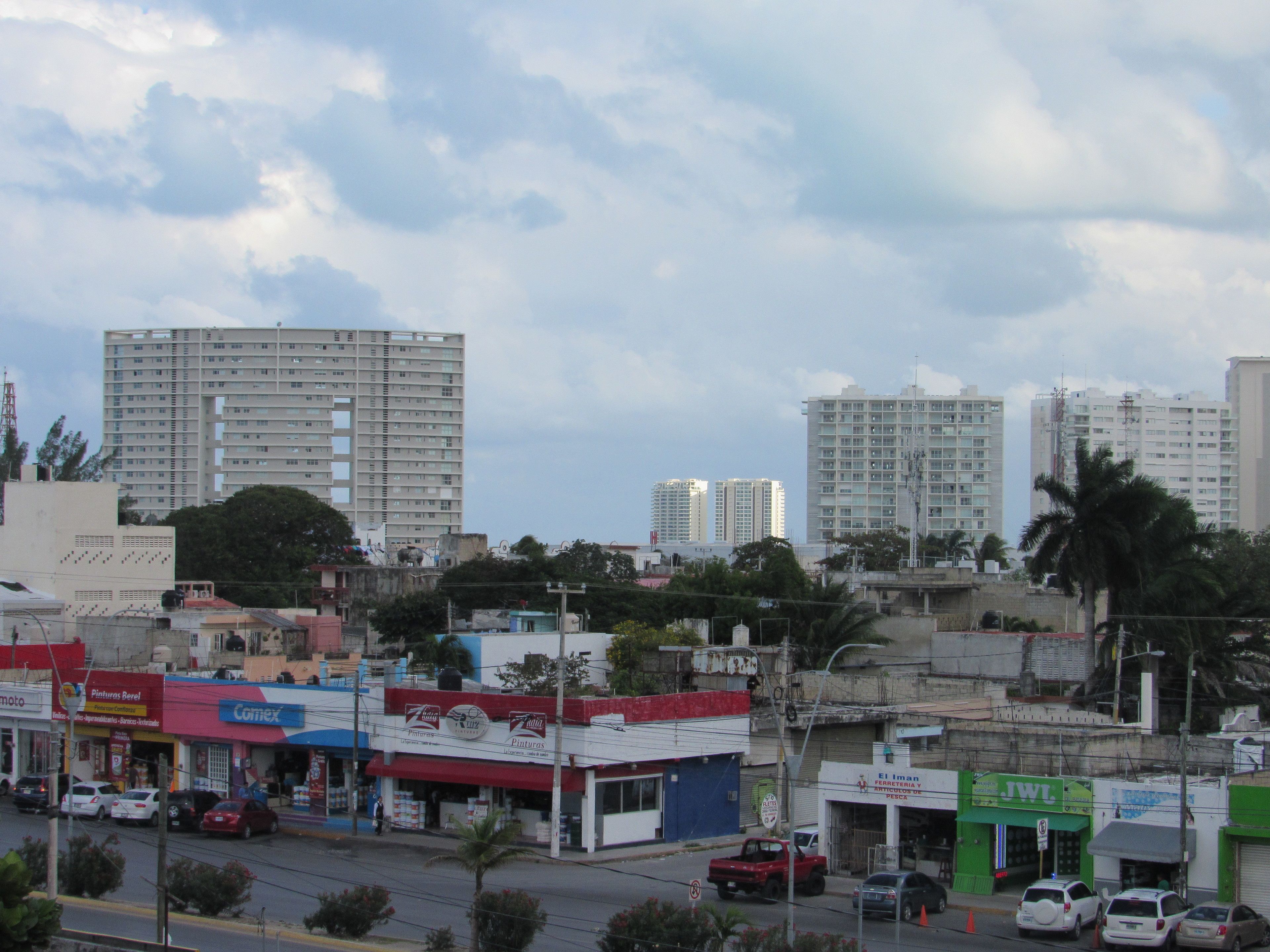 A cityscape with multiple-story buildings, a street with parked cars, and various storefronts.