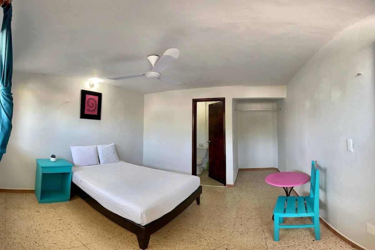 A bedroom with a bed, a ceiling fan, a small table, a chair, and a door leading to a bathroom.