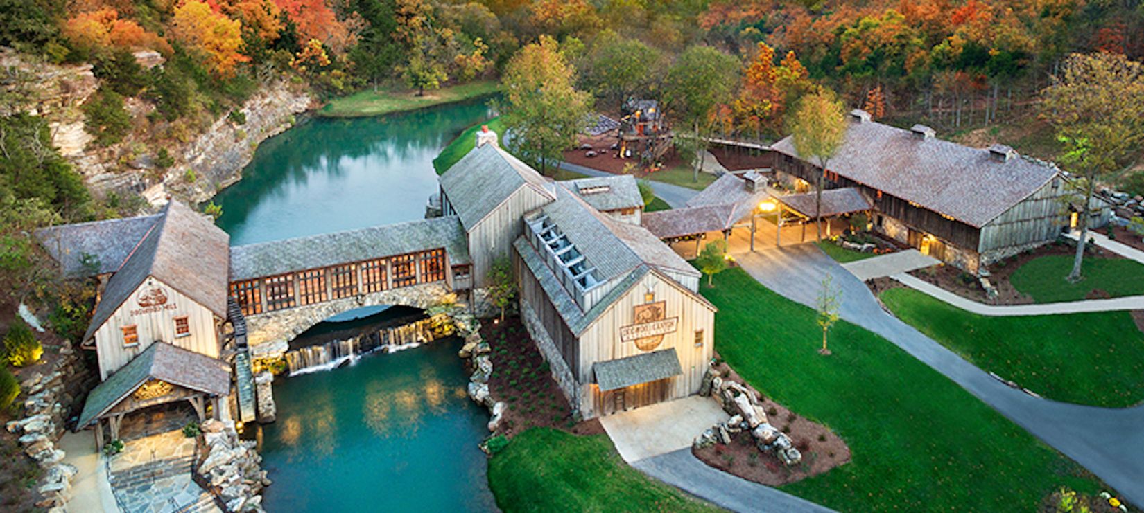 Un chalet rustique avec un pont de pierre enjambant un étang, le tout entouré d’un feuillage automnal.