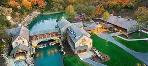 A rustic lodge with a stone bridge over a pond, surrounded by autumn foliage.