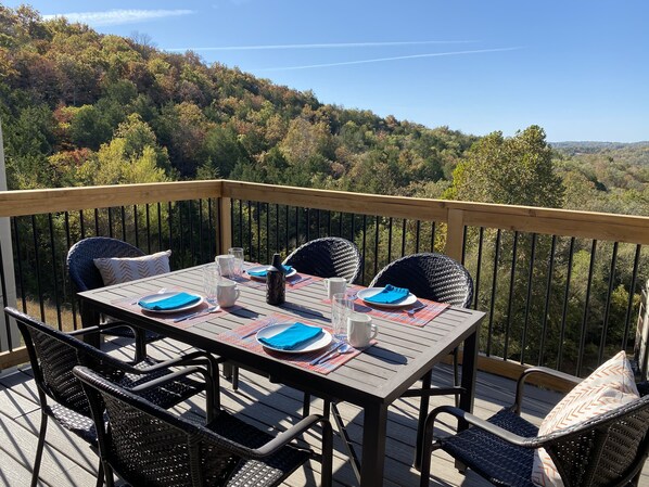 A deck with a table set for four, surrounded by wicker chairs, overlooking a forested hillside.