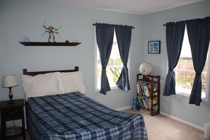 Un dormitorio con cama, mesita de noche, estantería y dos ventanas con cortinas.
