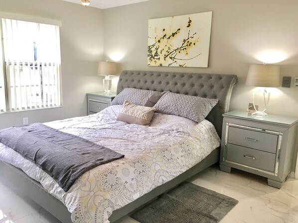 A bedroom with a large bed, two bedside tables, a nightstand, a wall-mounted lamp, and a window with blinds.