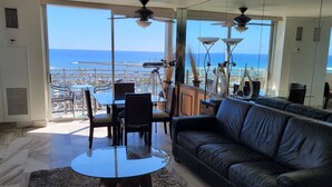 A living room with a leather sofa, a glass coffee table, and a dining table with chairs. There are ceiling fans and a view of the ocean through large windows. - Absolutely Oceanfront - Amazing view- Full Kitchen- full disability access. (honolulu)