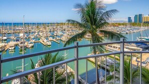 A marina with numerous boats docked, a palm tree in the foreground, and a city skyline in the background. - Absolutely Oceanfront - Amazing view- Full Kitchen- full disability access. (honolulu)