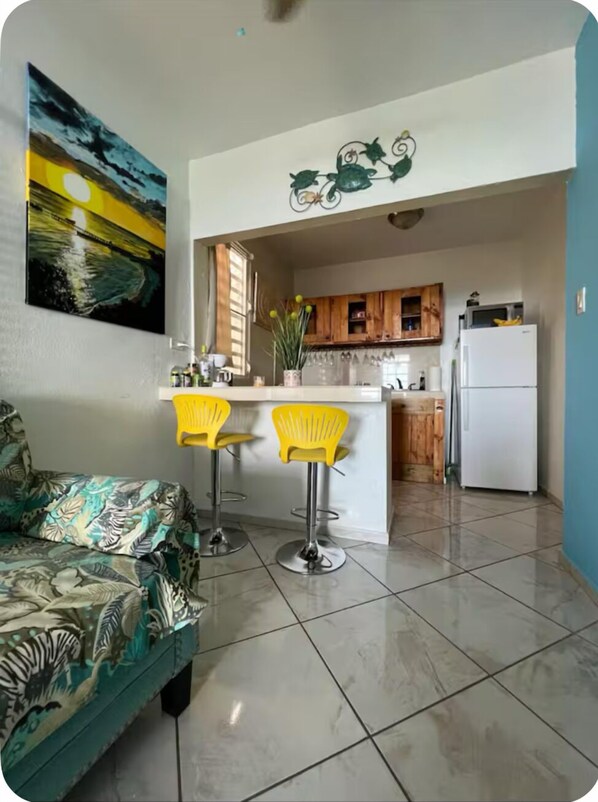 A compact living space with a sofa, two yellow bar stools, a kitchen with wooden cabinets, a refrigerator, and a painting on the wall.