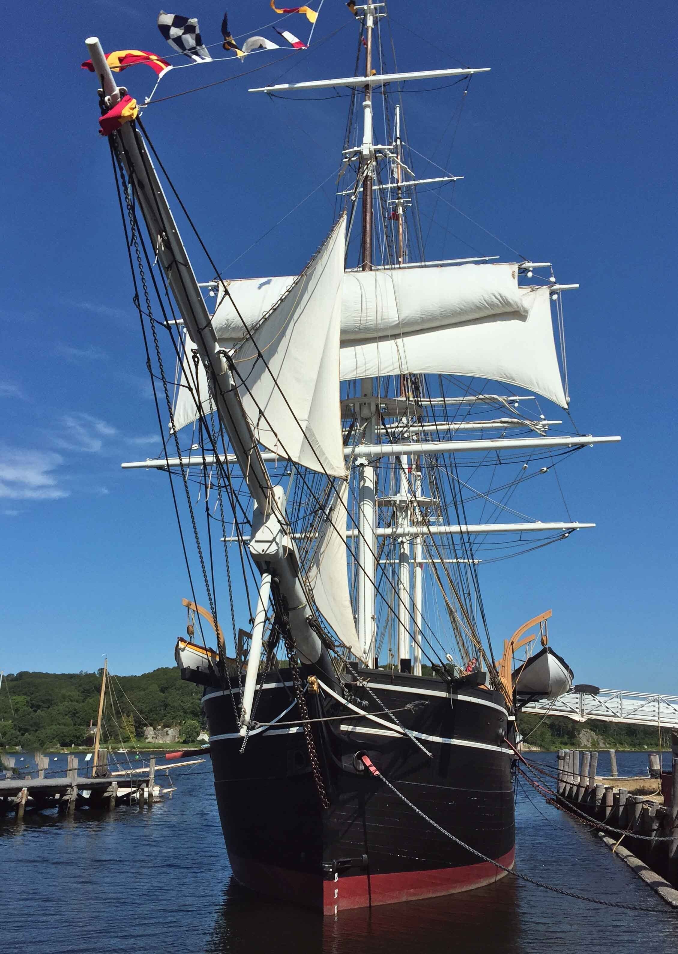 Un grand voilier était amarré à un quai, ses nombreux mâts et ses voiles se dressant vers le ciel, tandis qu’une eau calme l’entourait et que le ciel était dégagé.