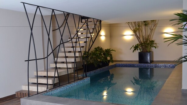 Piscina interior moderna com estrutura de vidro geométrica, escada com corrimão de metal e plantas em vasos.