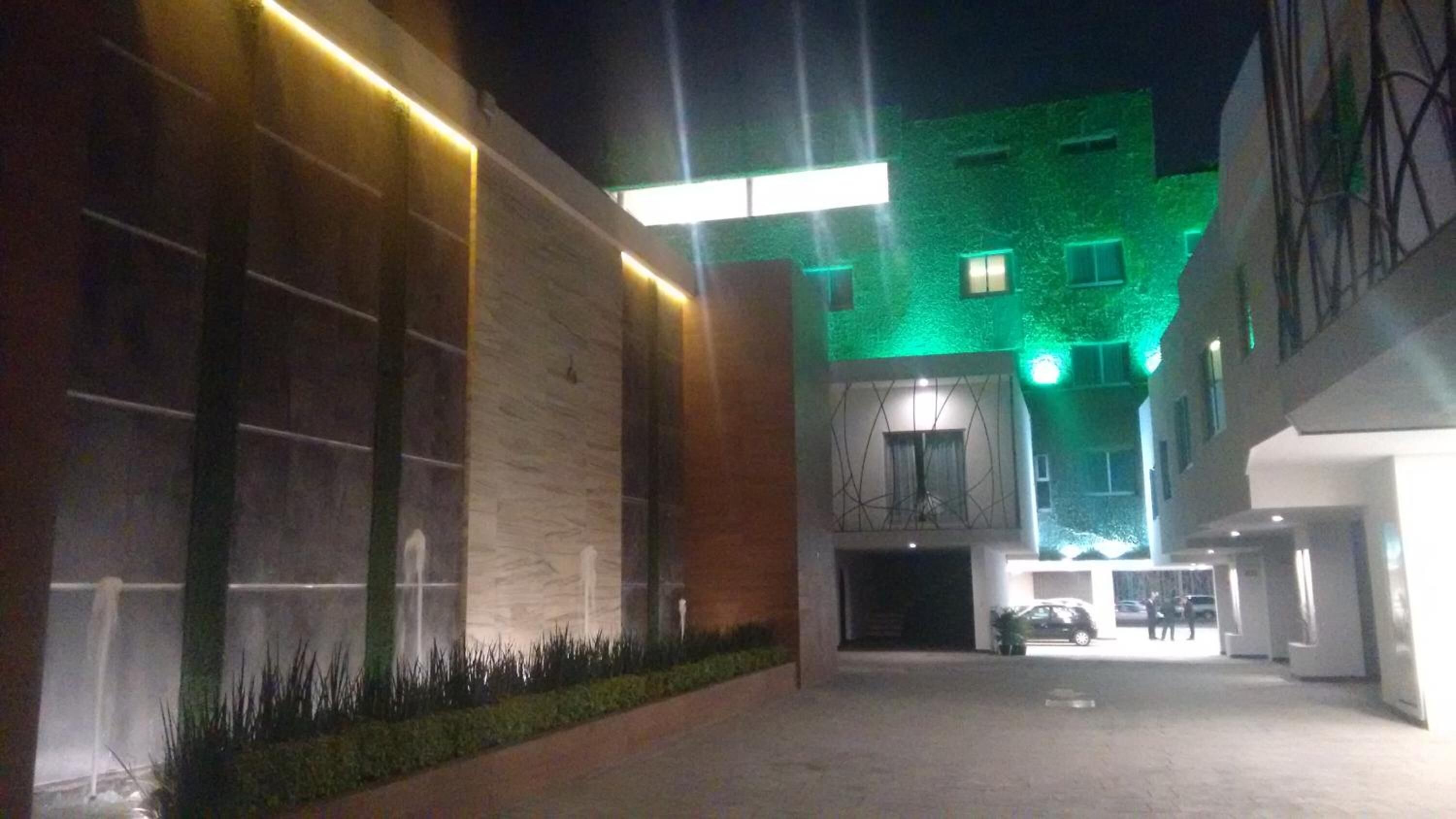 A modern building with a green-lit facade, a covered entrance, and a parking area.