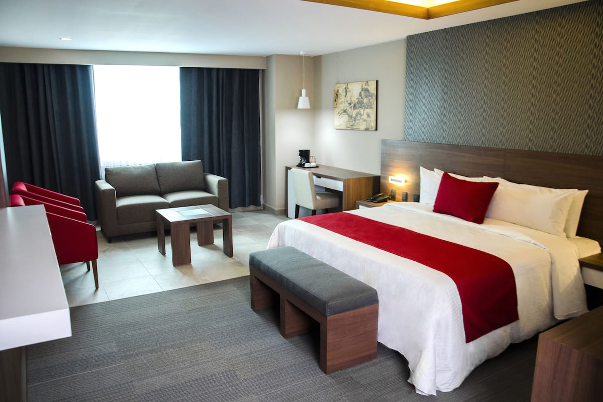 Une chambre d’hôtel avec un lit, un bureau, une chaise, un banc et une fenêtre avec des rideaux.