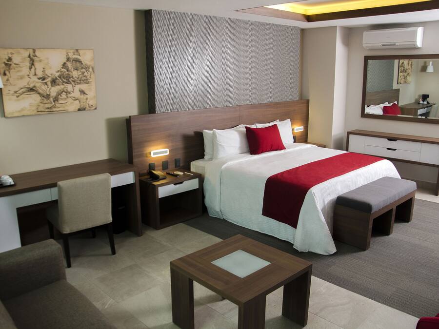 A hotel room with a bed, bedside tables, a desk, a chair, a small table, and a large mirror.