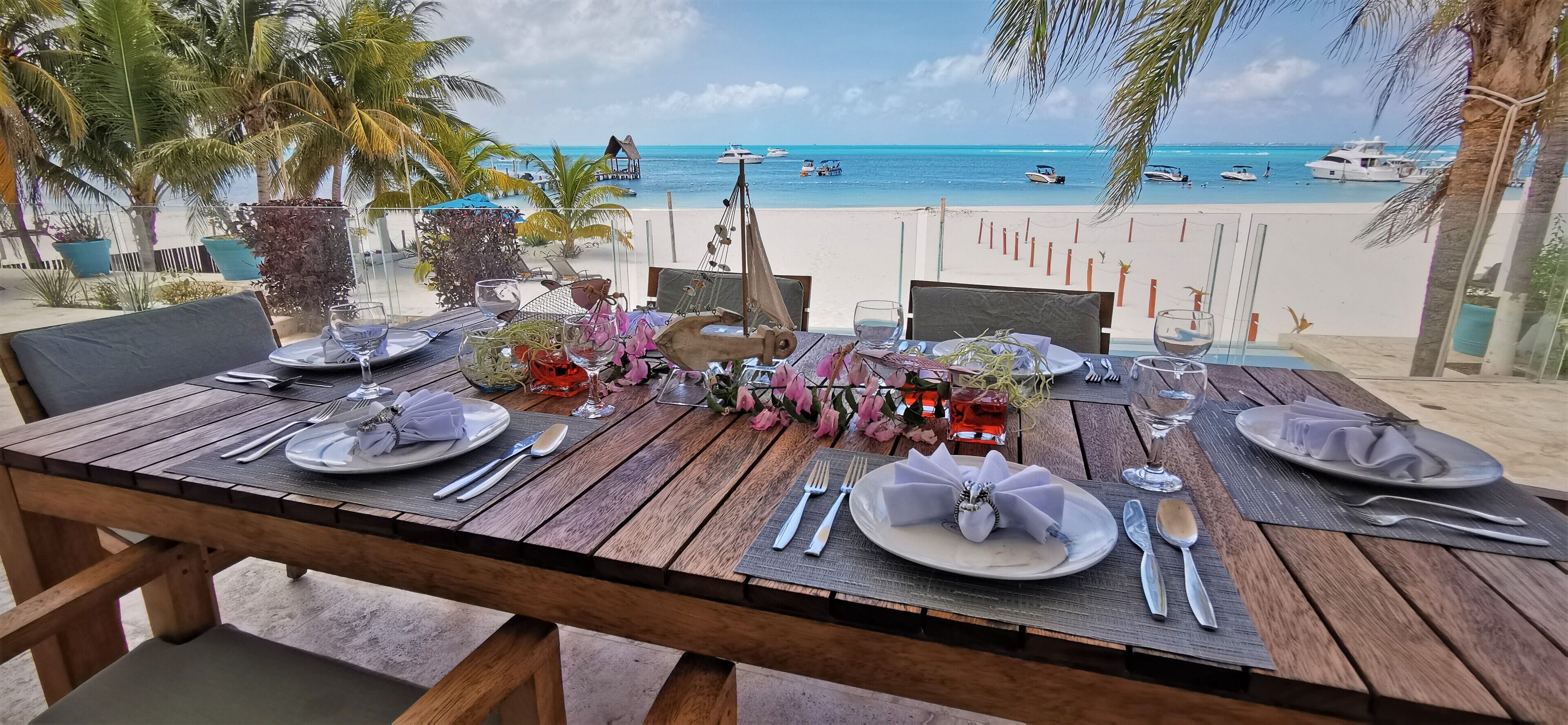 A wooden table set for two with plates, cutlery, and a vase of flowers, overlooking a beach with boats and palm trees.