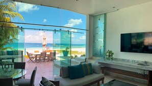 A modern living room with a glass wall overlooking a beach, furnished with a sofa, chairs, and a coffee table. - Exclusive & Safe Location: Turquoise Sea w/ White Sand Beach NO SEAWEED (Cancún)