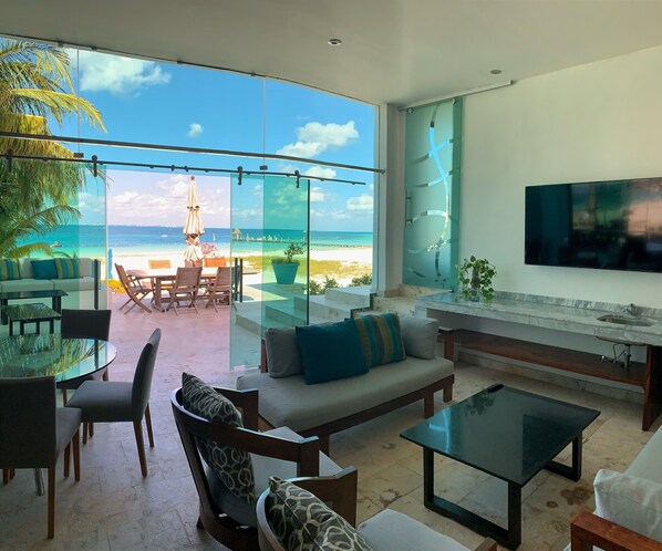 A modern living room with a glass wall overlooking a beach, furnished with a sofa, chairs, and a coffee table.