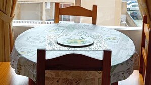 Une table ronde recouverte d’une nappe à motifs, des chaises en bois et une fenêtre avec des stores.