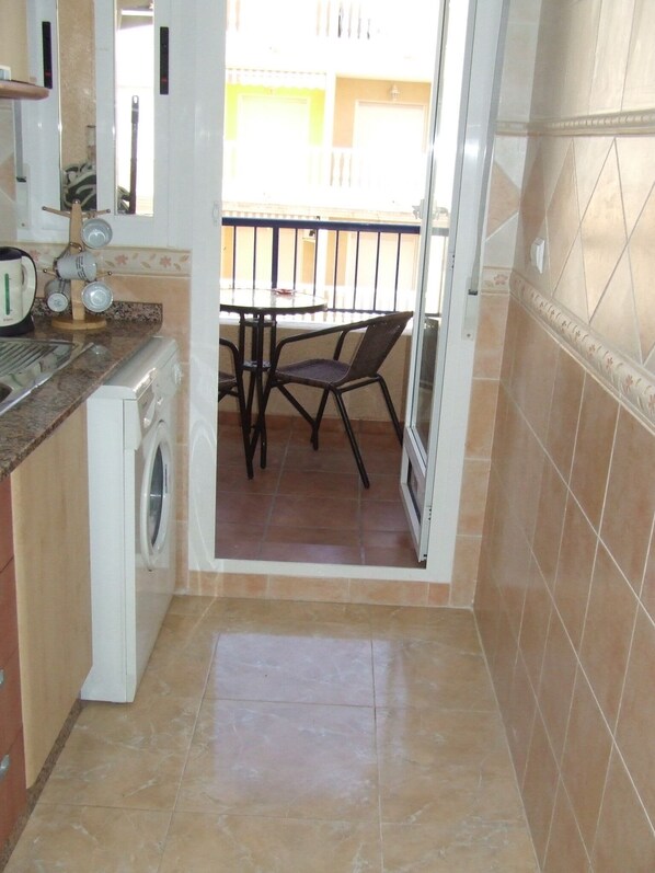 A compact kitchen with a washing machine, a dining table with chairs, and a view of a balcony.