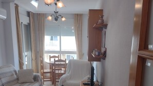 Un salon confortable avec un canapé, une table à manger et une chaise. On y trouve une petite table d’appoint en bois et une lampe. Un luminaire est visible au plafond.