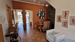 Un vaste espace de vie doté d’un plafond haut, d’un mur en pierre servant de décor et d’une table à manger avec des chaises. Des œuvres d’art encadrées ornent le mur, et une machine à coudre est posée sur une table.