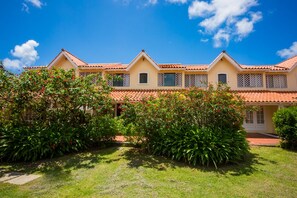 A two-story building with a tiled roof and multiple windows, surrounded by lush greenery and a clear blue sky.