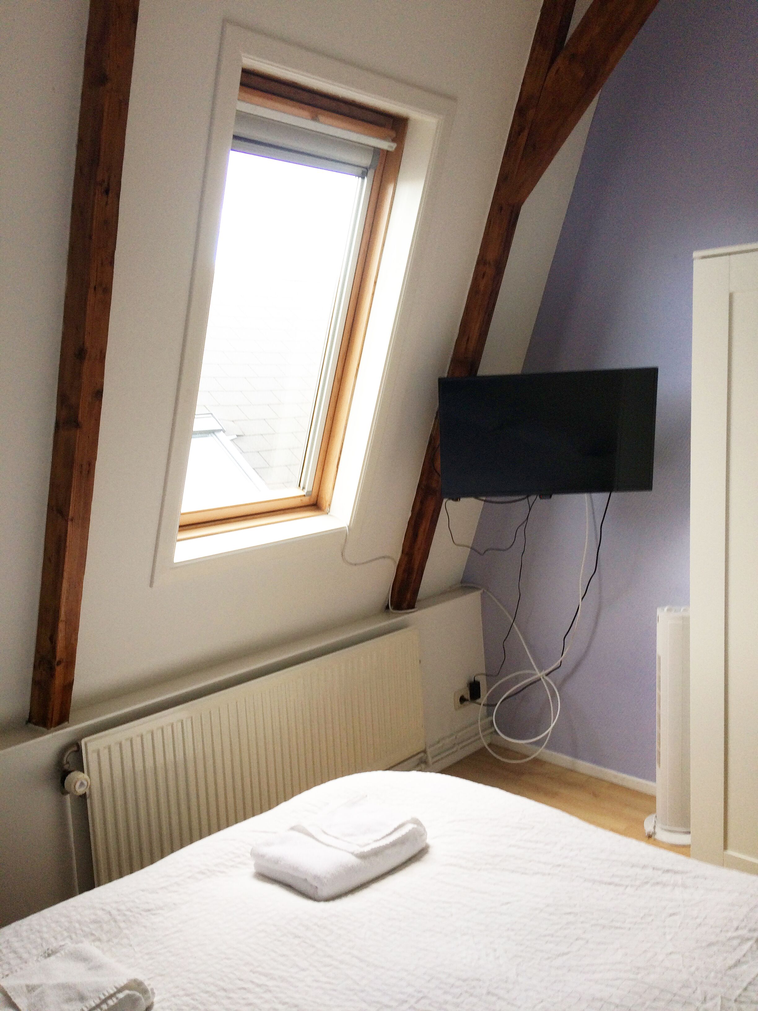 Un dormitorio con techo inclinado, ventana con persianas, televisor de pantalla plana montado en la pared y radiador debajo de la ventana.