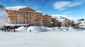 Lumipeitteinen hiihtokeskus, jossa on useita kerroksia sisältäviä puurakennuksia, hiihtohissejä ja kirkas, sininen taivas.
