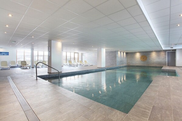 A large indoor swimming pool with clear water, surrounded by lounge chairs and a tiled floor. - Chalet in Paradiski with Spa & Pool Access (Plagne Villages)