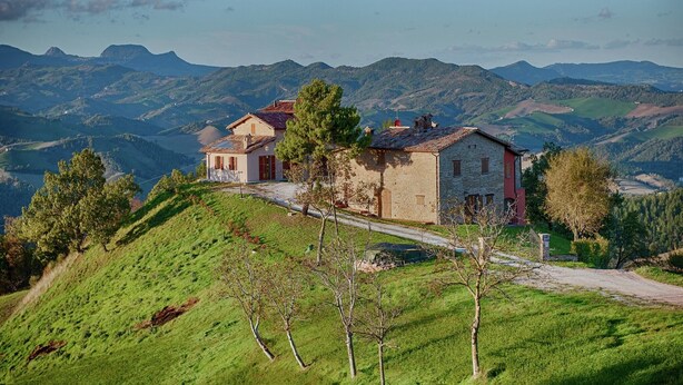 Une maison avec une façade en pierre, un toit rouge et une vue sur des collines verdoyantes à l’arrière-plan.