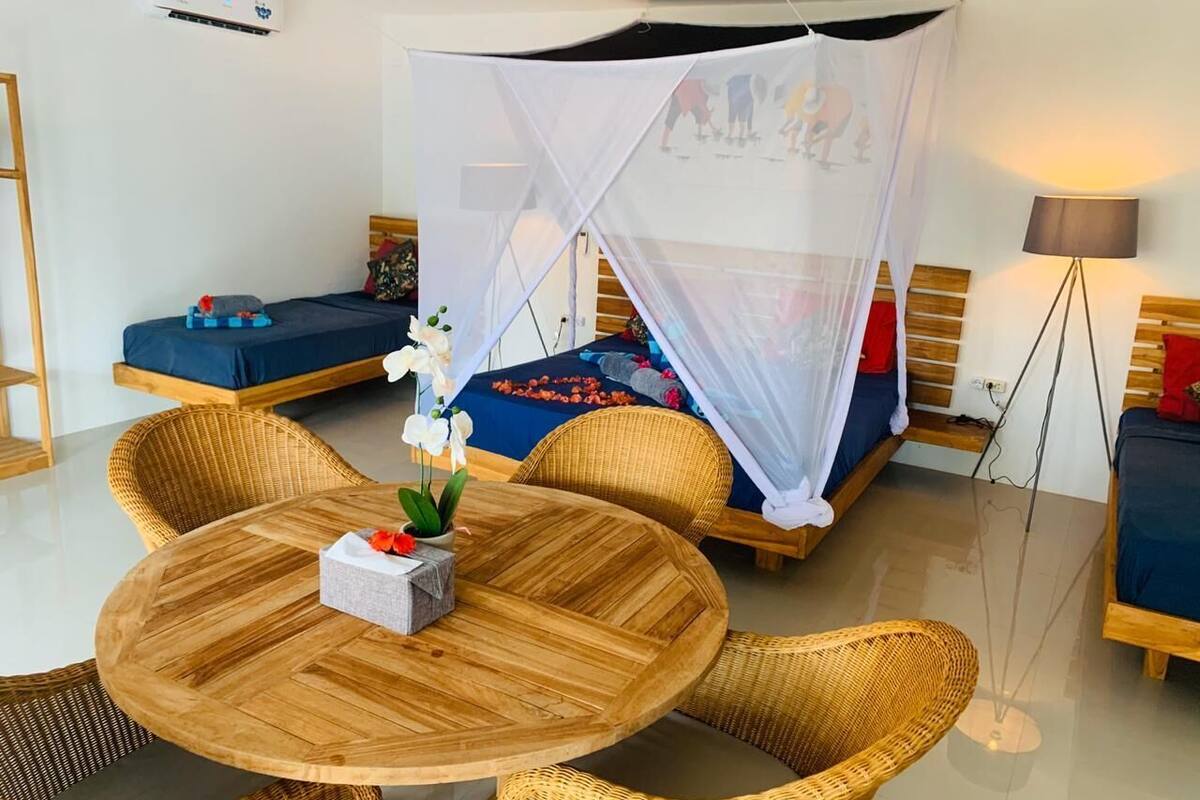 A room with a round wooden table, wicker chairs, a bed with a canopy, and a bedside lamp.