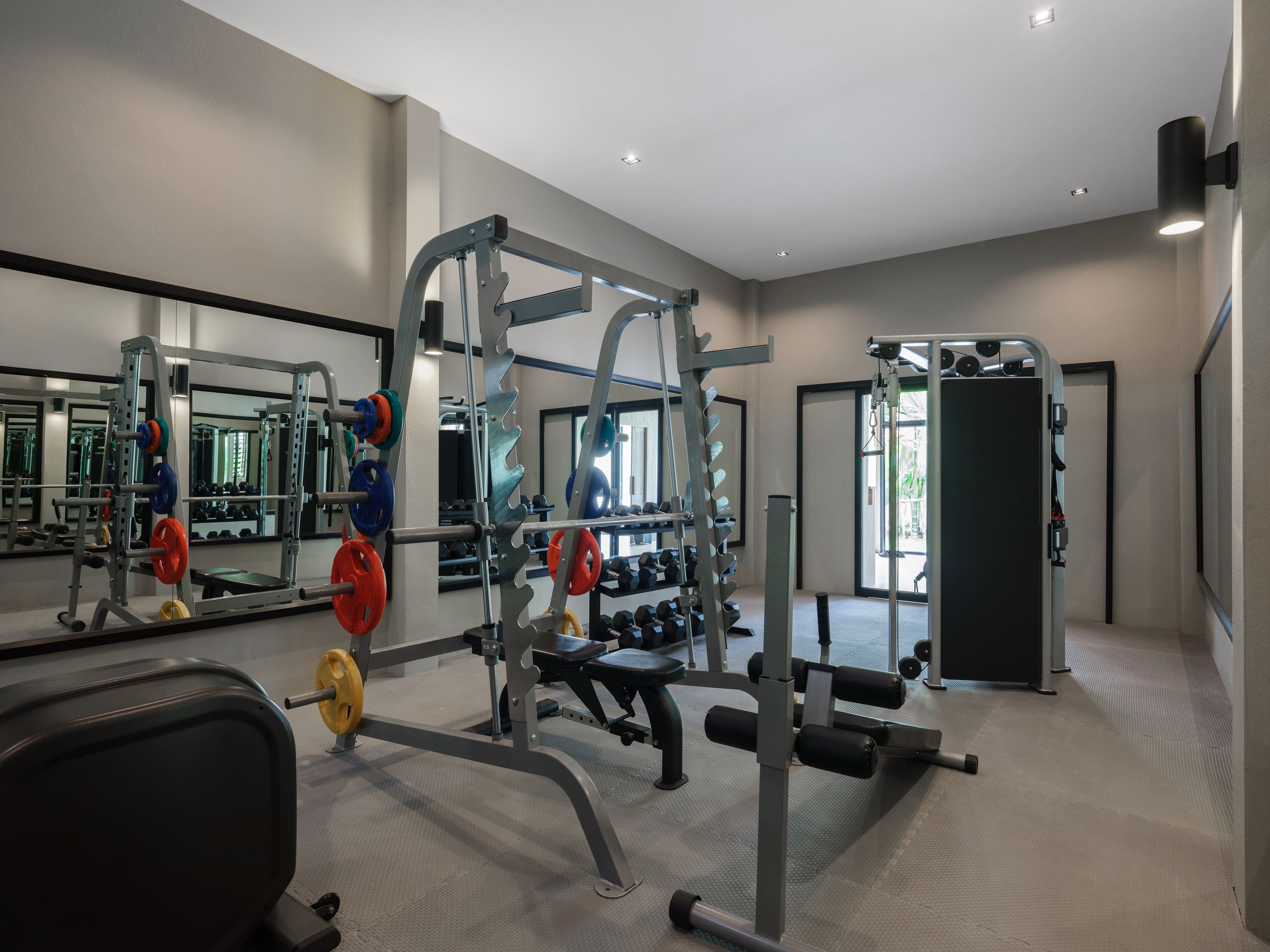 A modern gym with various weightlifting equipment, mirrors, and a large window providing natural light.