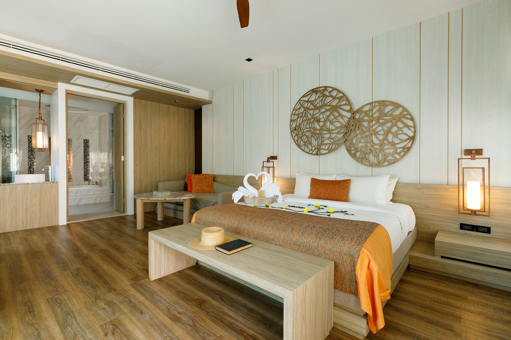 A modern hotel room with a wooden floor, a bed with a white and orange bedspread, a bedside table with a lamp, and a wall with decorative circular patterns.