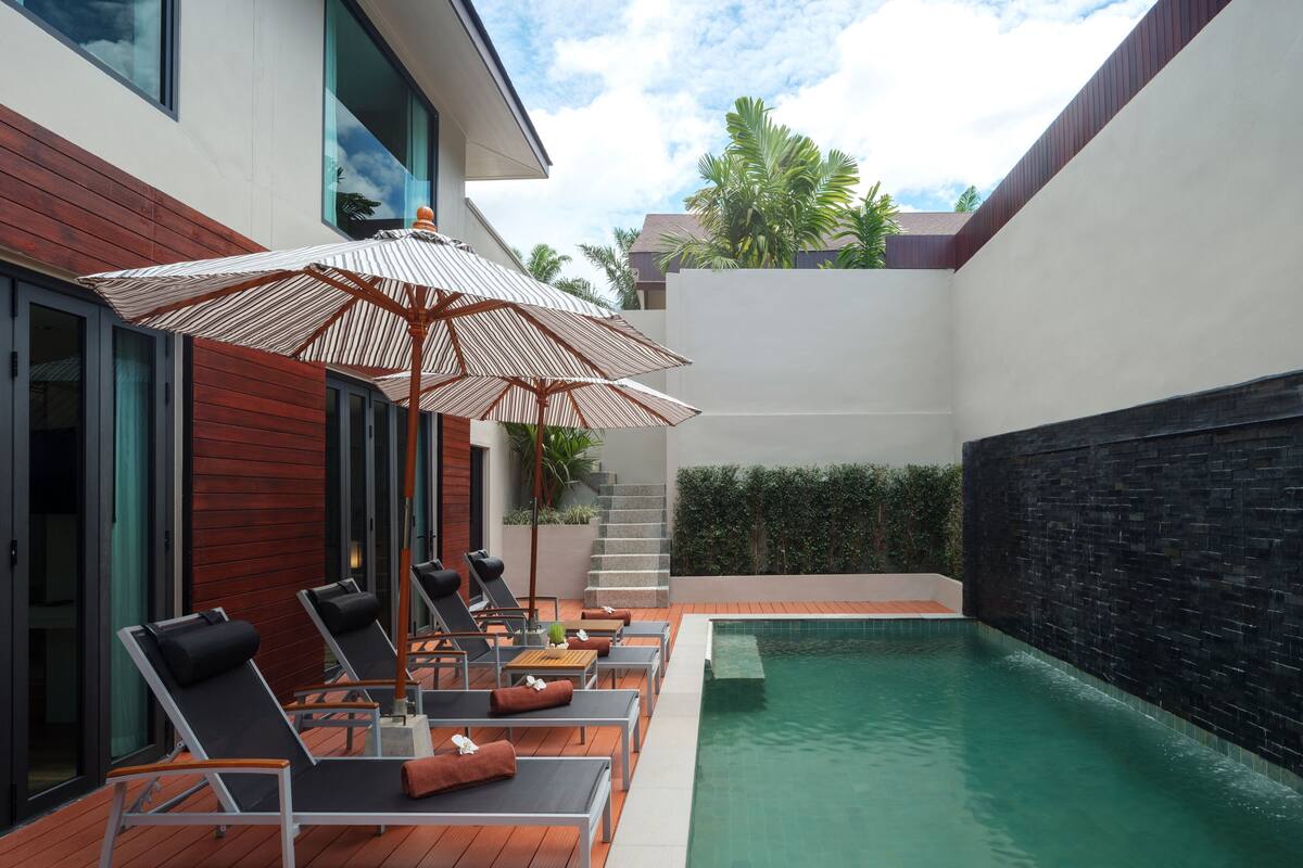 A modern outdoor pool area with lounge chairs and umbrellas, a wooden deck, and a stone wall.