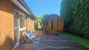 Una terraza de madera con una silla de descanso, un barril y una casa de ladrillo.