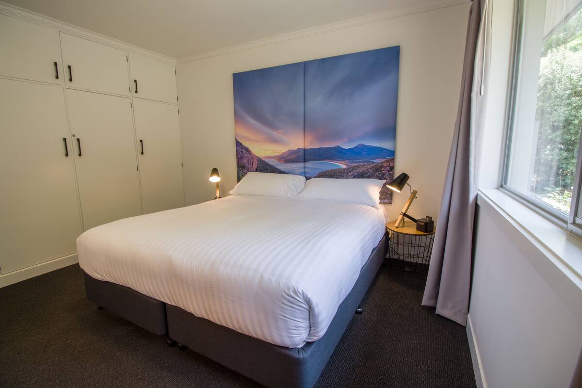 Un lit bien fait dans une chambre d’hôtel, avec un grand tableau représentant un paysage accroché au mur, une table de chevet avec une lampe et une fenêtre avec des rideaux.