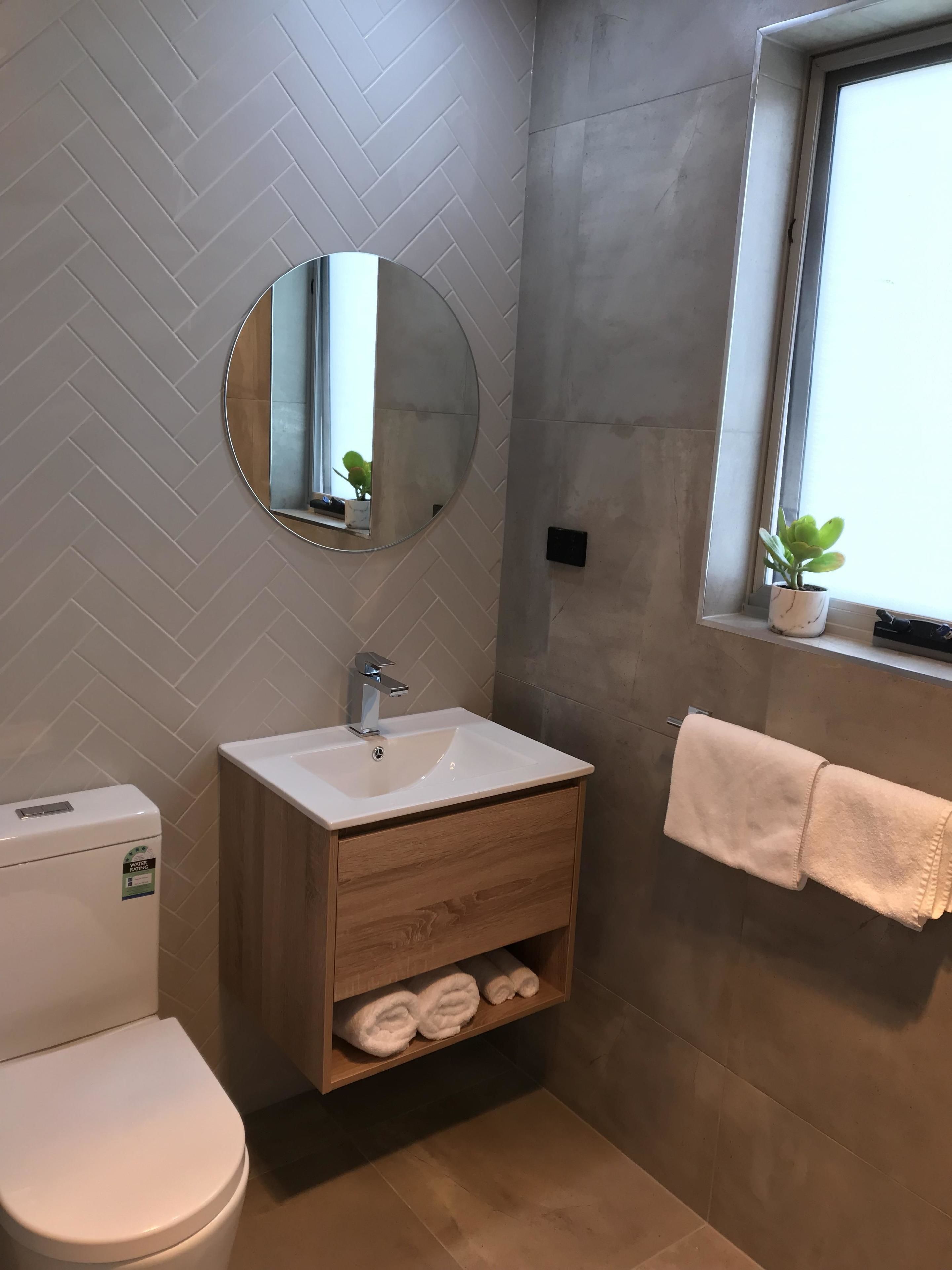 A bathroom with a white sink, wooden vanity, a round mirror, a towel rack, and a potted plant by the window.