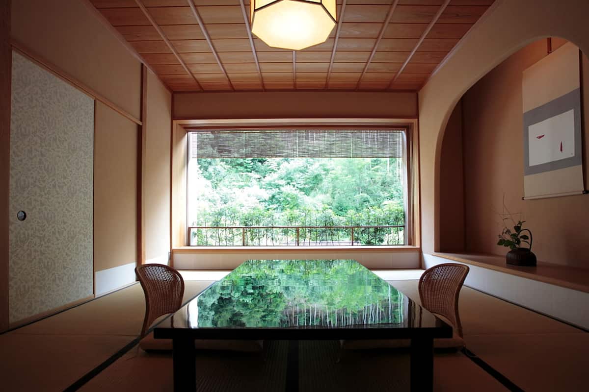 A traditional Japanese room with tatami flooring, a low table, two chairs, a large window with a view of greenery, and a ceiling light fixture.