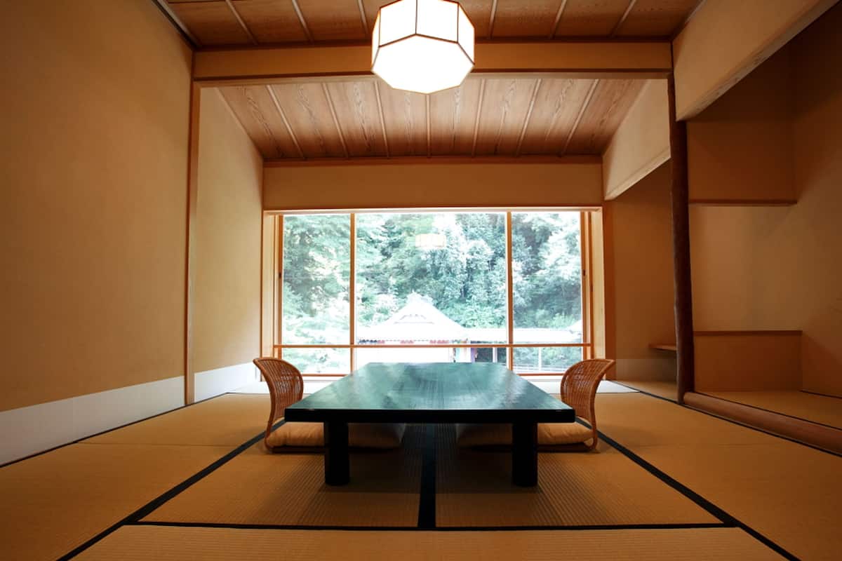 A traditional Japanese room with tatami flooring, a low table, and a large window offering a view of trees.