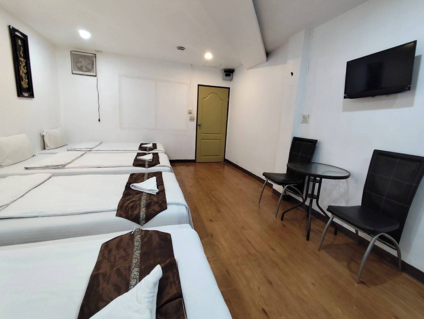 Une chambre d’hôtel avec trois lits simples, une petite table avec deux chaises, un téléviseur fixé au mur et une porte avec un interrupteur.