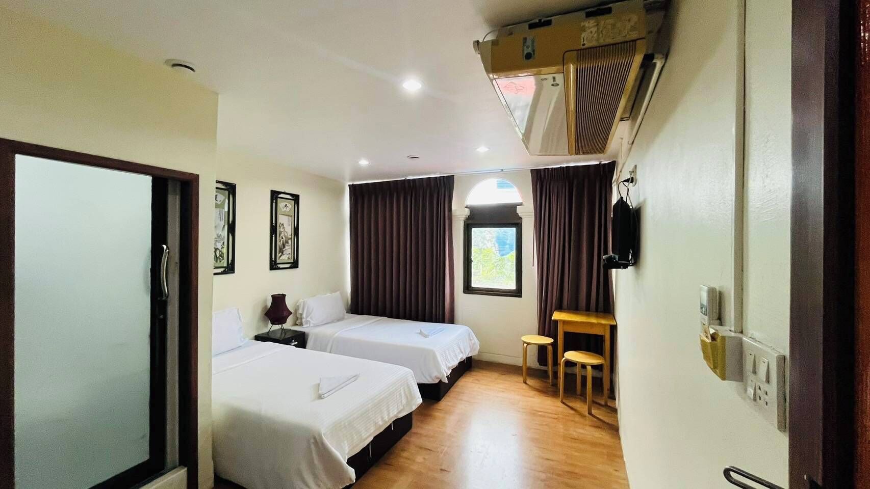 Een hotelkamer met twee bedden, een klein tafeltje, een stoel, een raam met gordijnen en een plafondventilator.