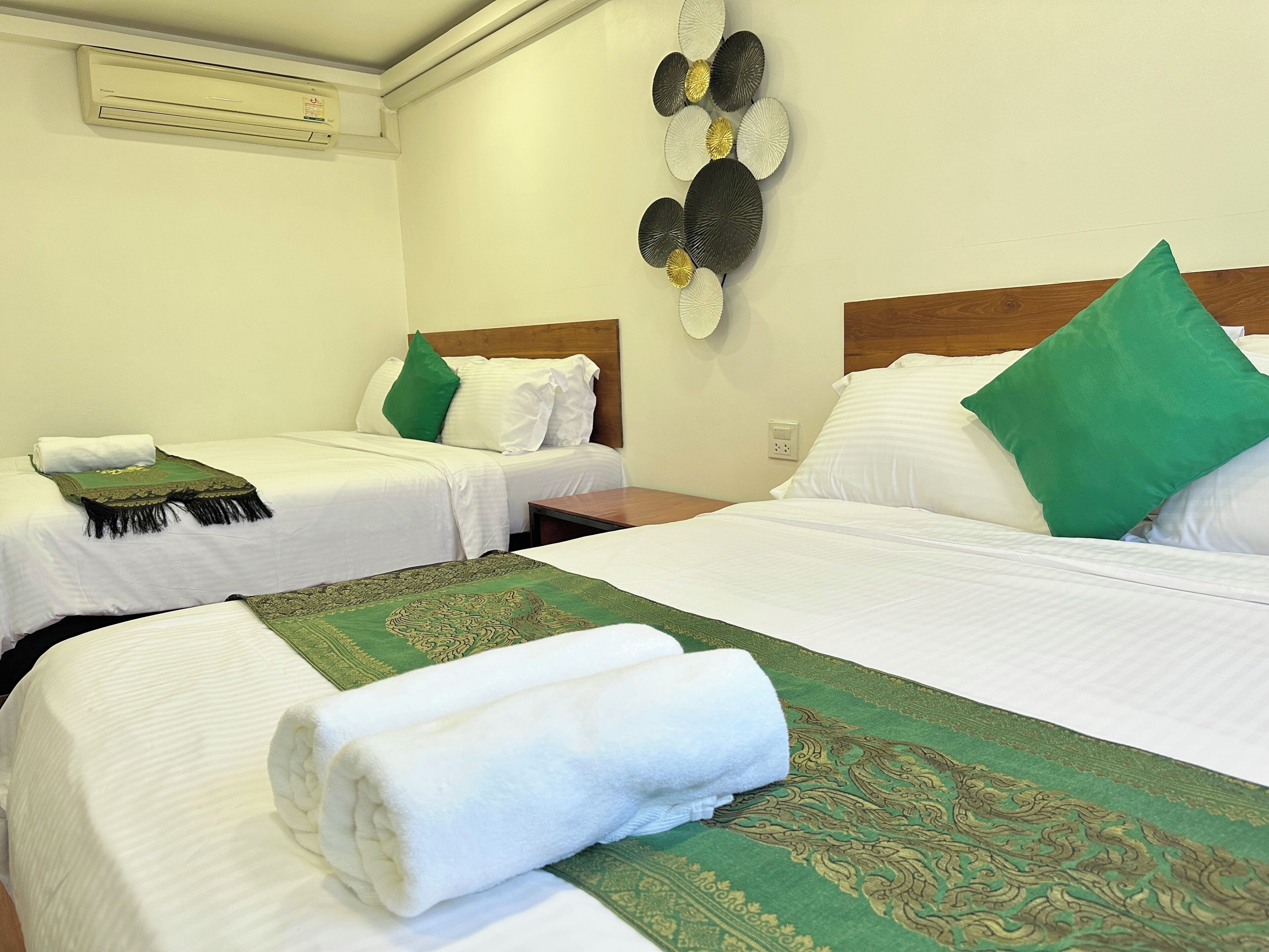 A hotel room with two beds, each with white linens and green pillows. There is a decorative wall piece above the beds and an air conditioning unit on the wall.