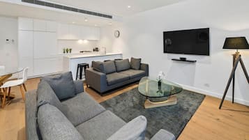 Uma sala de estar moderna com um sofá cinzento, uma mesa de centro de vidro e uma televisão de tela plana na parede.