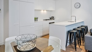 A modern kitchen with white cabinetry, a central island with stools, a clock on the wall, and a dining area with a geometric glass bowl on the table. - Anchorage Apartments Hampton (Hampton)