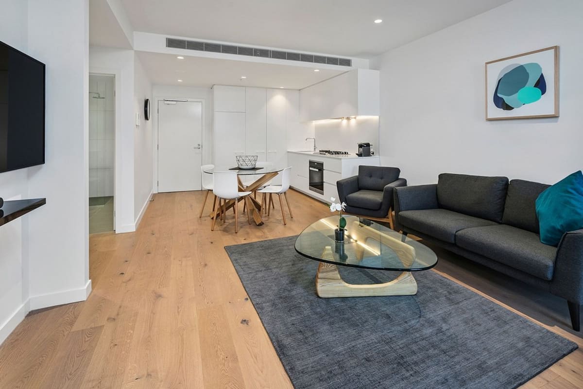 A modern living room with a glass coffee table, grey sofa, and a dining area with white chairs.