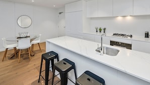 A modern kitchen with white cabinetry, a marble countertop, a sink, and a stove. There is a dining area with a round table and chairs. - Anchorage Apartments Hampton (Hampton)