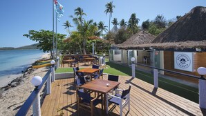 Una terraza de madera con mesas y sillas da vista a una playa, un edificio con techo de paja y un asta con una bandera.