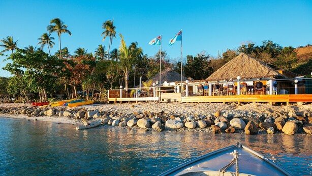 Un hotel frente a la playa con una estructura de techo de paja, una costa rocosa y varias kayaks.