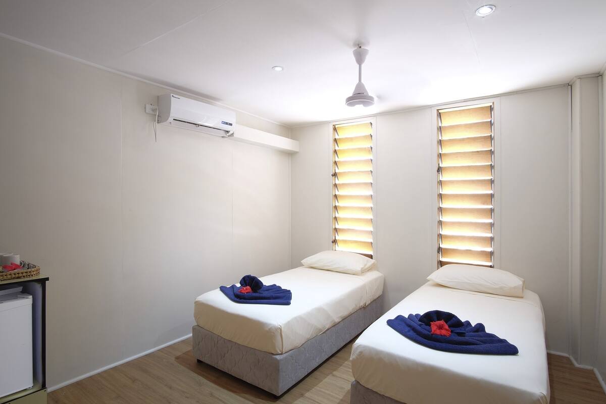 A room with two single beds, a ceiling fan, an air conditioning unit, and a small table with a basket.
